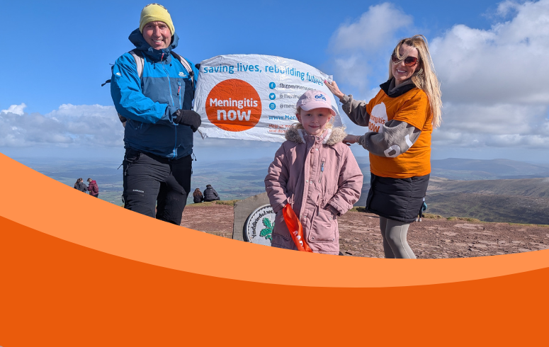 Young girl in pink hat and coat stands in front of a banner held by her parents at the top of a mountain
