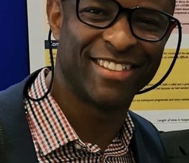 A man wearing black glasses and a checkered shirt smiles to the camera.