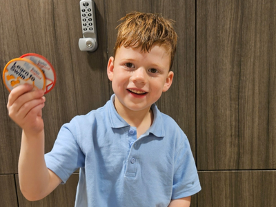 A smiling young boy in a light blue shirt holds up swimming badges proudly. The background features a wooden locker.