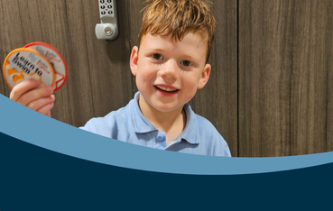 White child with auburn hair stands in front of wooden cabinet holding two swimming badges