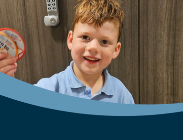 White child with auburn hair stands in front of wooden cabinet holding two swimming badges