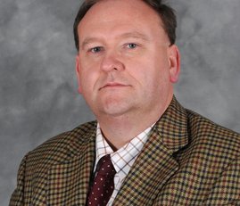 A man wearing formal clothes of a blazer and shirt against a greyish background.