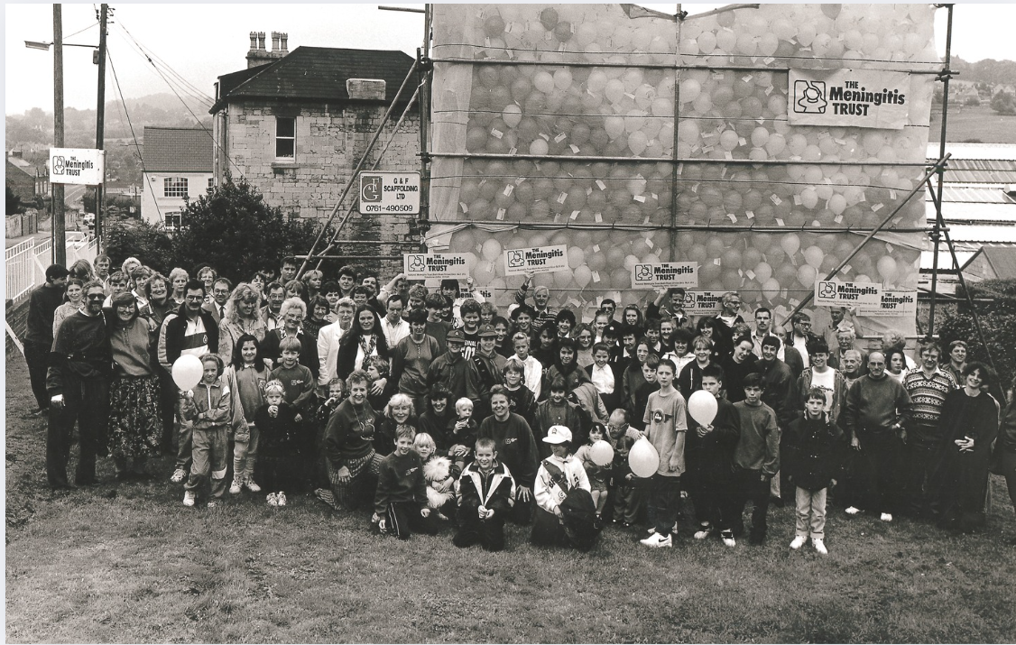 A large group of adults and children, some holding balloons, pose outside in front of a building with scaffolding. Overlay text reads "40 years saving lives, rebuilding futures," with a "Meningitis Now" logo.