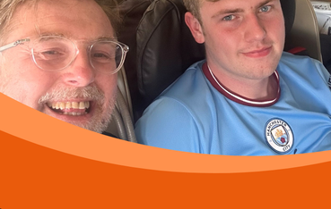 Two men, one in foreground with glasses and beard, and one in background with blonde hair wearing a Manchester City football t-shirt, smile at camera