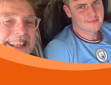 Two men, one in foreground with glasses and beard, and one in background with blonde hair wearing a Manchester City football t-shirt, smile at camera