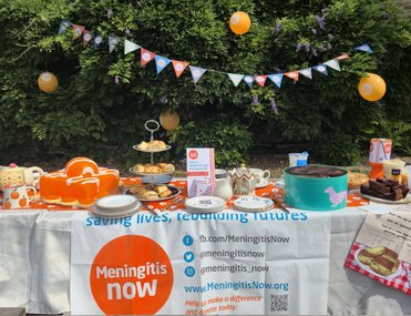 A table in the garden of orange themed baked goods with decorations and a collection of money for Meningitis Now