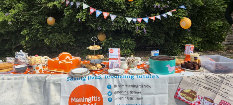 A table in the garden of orange themed baked goods with decorations and a collection of money for Meningitis Now