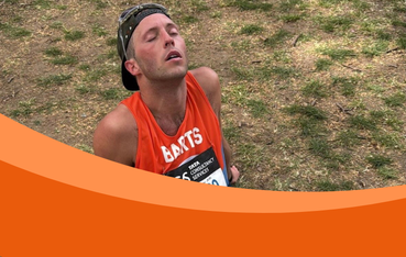 White man sits on ground with eyes closed and wearing an orange running vest, a baseball cap on backwards and sunglasses on his head