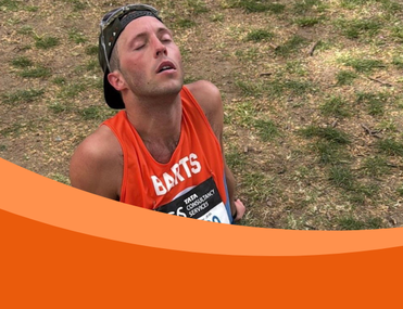 White man sits on ground with eyes closed and wearing an orange running vest, a baseball cap on backwards and sunglasses on his head