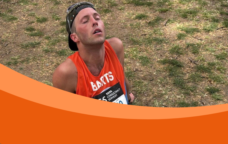 White man sits on ground with eyes closed and wearing an orange running vest, a baseball cap on backwards and sunglasses on his head