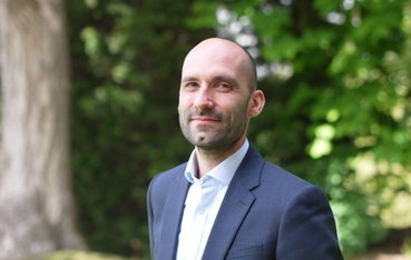 A white man with stubble in a suit smiling at the camera, set in a garden.