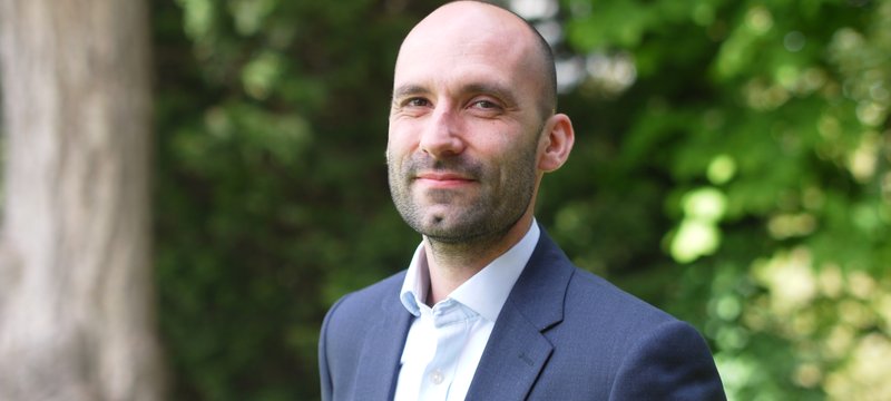 A white man with stubble in a suit smiling at the camera, set in a garden.