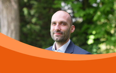 A white man with a stubble dress in a suit looking at the camera. The background is in a garden.