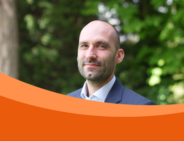 A white man with a stubble dress in a suit looking at the camera. The background is in a garden.