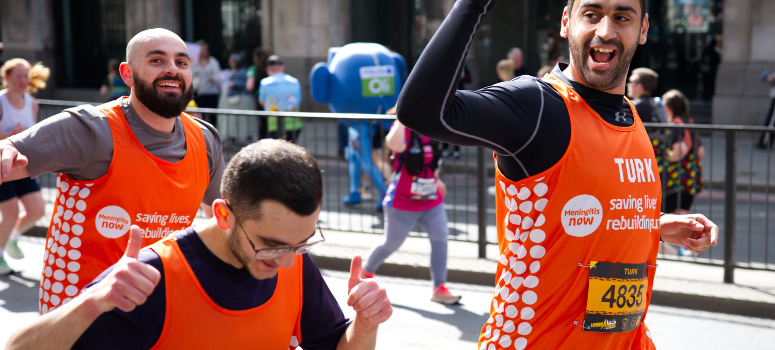 Three people in orange running vests taking part in a running event for Meningitis Now. They are cheering and are excited.