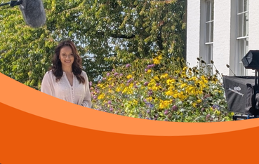 Woman TV presenter stands in front of white building amongst flowers with sound boom and film camera in shot