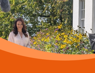 Woman TV presenter stands in front of white building amongst flowers with sound boom and film camera in shot