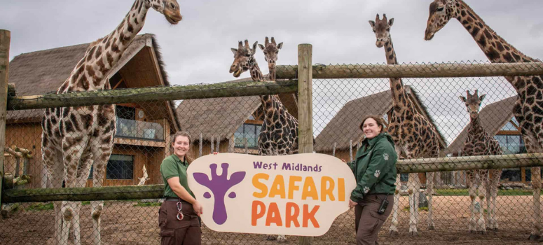 Two zoo keepers holding a sign for West Midlands Safari Park in front of the giraffe enclosure