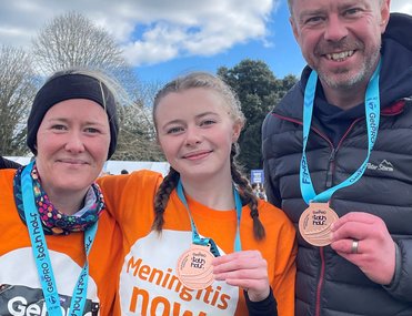 Three smiling individuals wearing orange "Meningitis Now" shirts and race numbers proudly display medals.