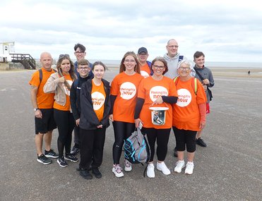A group of people wearing orange "Meningitis Now" shirts stand together on a beach promenade, smiling. The mood is cheerful and supportive.
